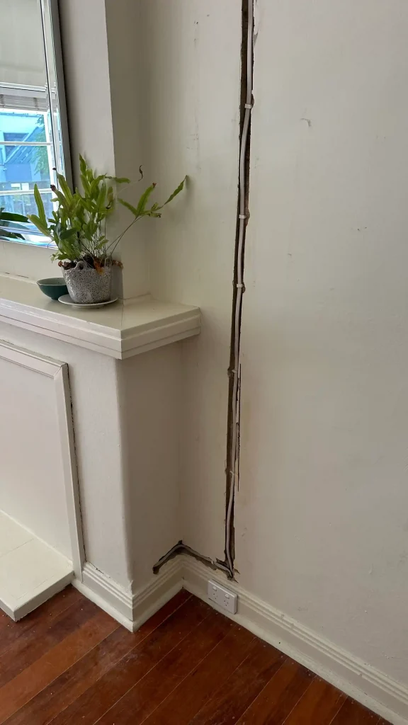 Narrow vertical wall damage exposing pipes and wiring in a white painted room.