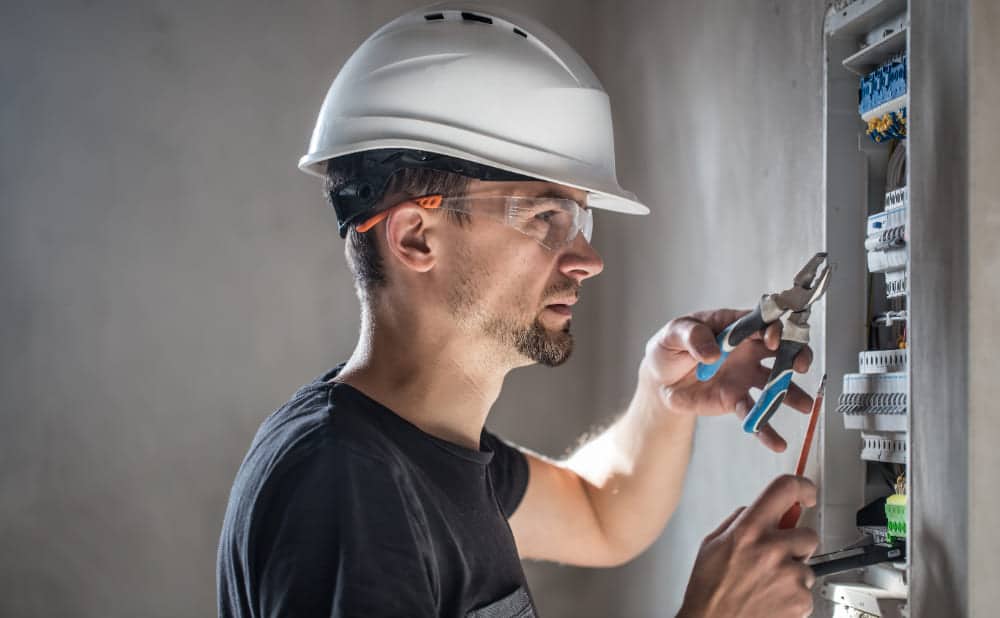 Electrician is working on electrical repairs in a panel board, wearing a safety helmet and protective glasses.