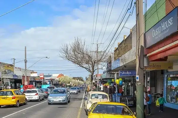 A vibrant street in Hampton, Victoria, featuring a variety of shops, parked cars, and pedestrians on a sunny day, with a mix of modern and vintage vehicles.