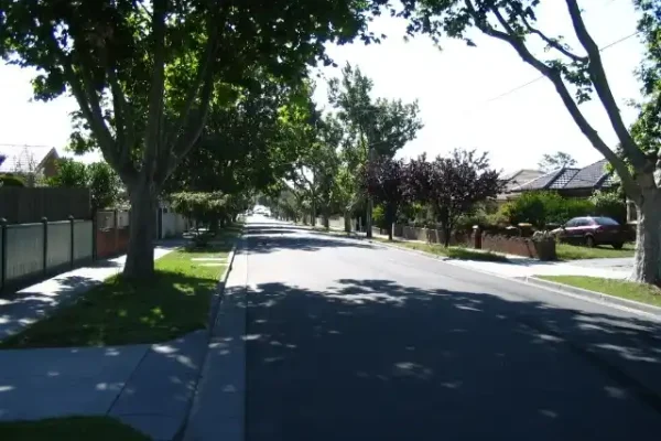 A tree-lined suburban street in Hughesdale with shaded sidewalks and residential houses under clear skies.